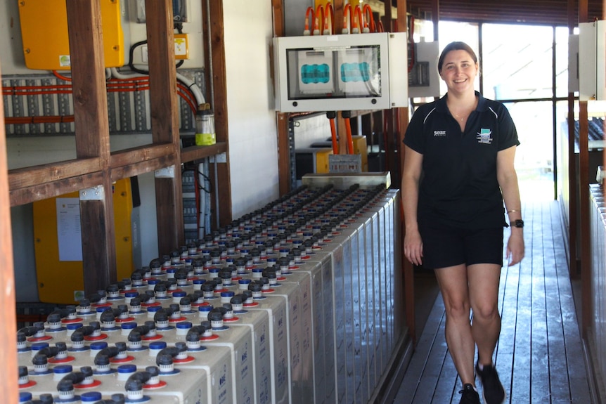 A woman stands beside a vast number of batteries inside a large shed.