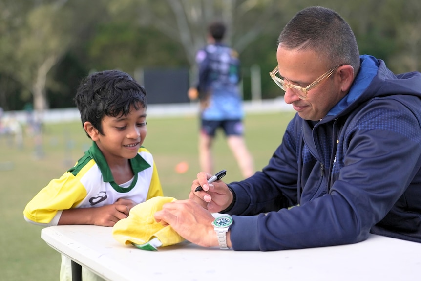 Usman Khawaja seated at a table outdoors wears glasses as he signs a cap for a young boy looking on smiling.