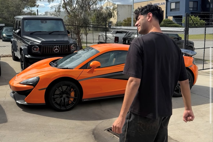 A man wearing black top with back to camera standing in front of orange luxury car