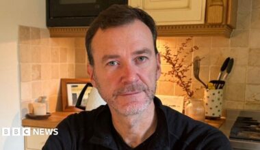 Mr Macleod is pictured sitting in his kitchen. He has short brown hair and a light grey beard. He is wearing a black half-zip top.