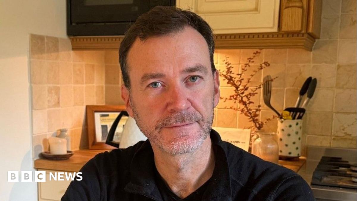 Mr Macleod is pictured sitting in his kitchen. He has short brown hair and a light grey beard. He is wearing a black half-zip top.