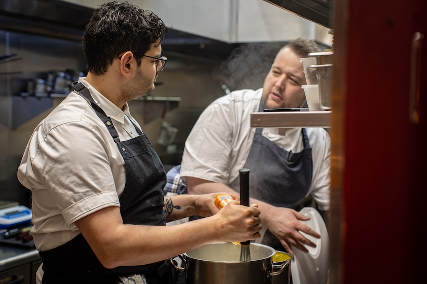 two men in white shirts and blue aprons talk over a pot on a stove in a kitchen