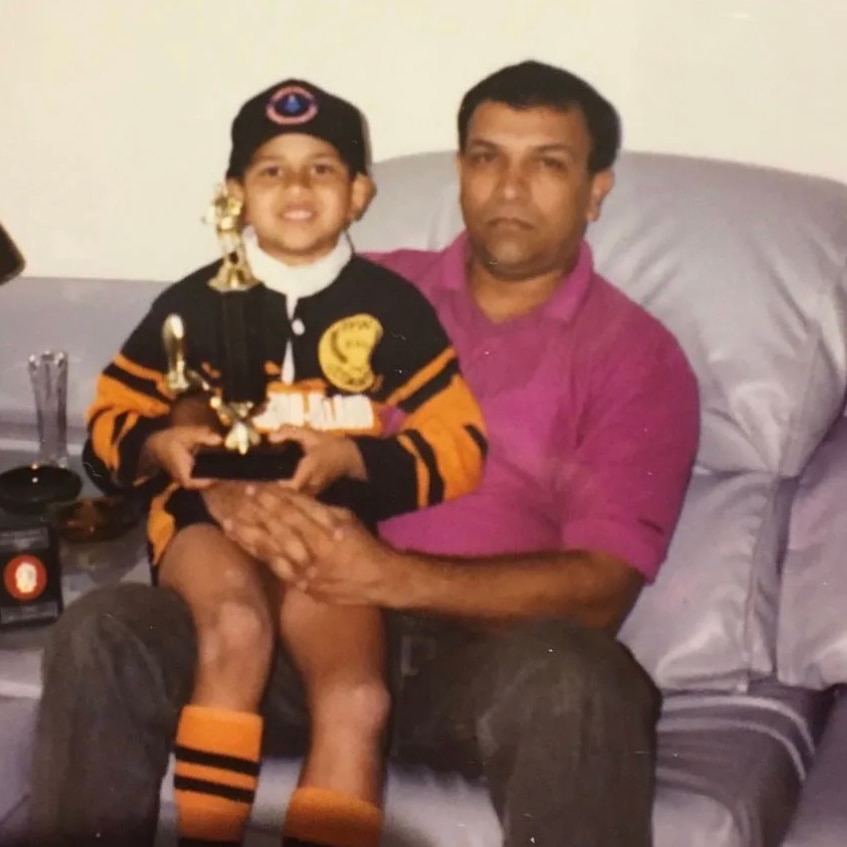 Usman Khawaja as boy sitting on his father's lap on couch. He holds several trophies. The boy is smiling while the father isn't.