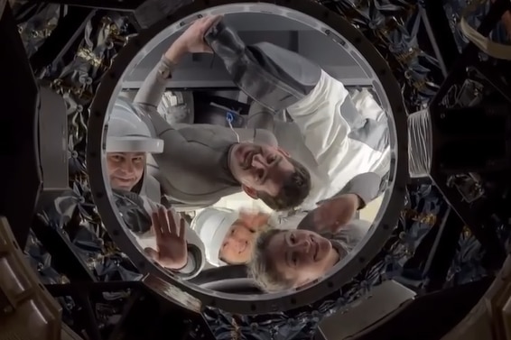 The faces of two male and female astronauts smiling and waving from inside a small circular opening in a space capsule hatch.