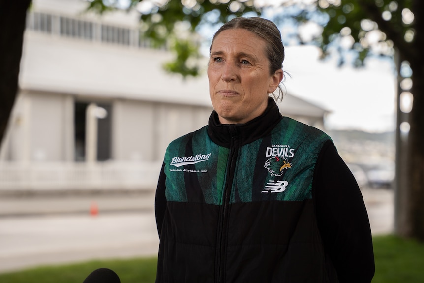 Kath McCann, in a Tasmania Devils jumper, addresses the media in a park.
