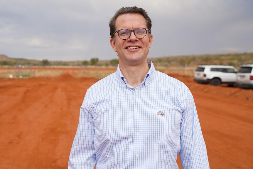 A white man in a blue plaid and white button up shirt, with brown hair, circle brown framed glasses, red dirt.