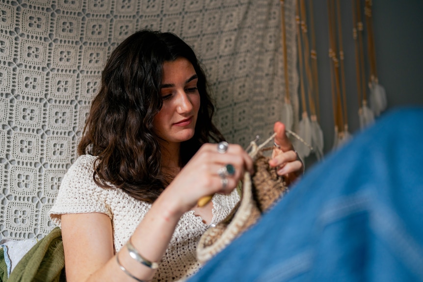 Close up of Lucy, wearing a crochet top and a green scarf, sitting on her bed crocheting a beanie with a soft smile.