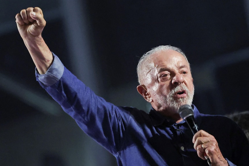 Luiz Inacio Lula de Silva in a denim blue shirt with the sleeves rolled up raising a fist while speaking into a microphone.