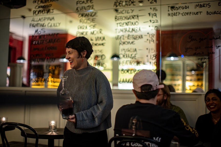 A woman laughs and walks across a restaurant holding a carafe of water with a big mirror behind and a wine list written on it