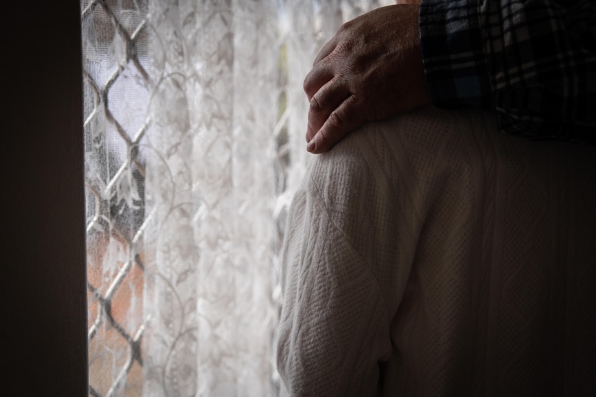 A man's left hand rests on top of a young girl's left shoulder, hugging her into him. They are looking through a window.