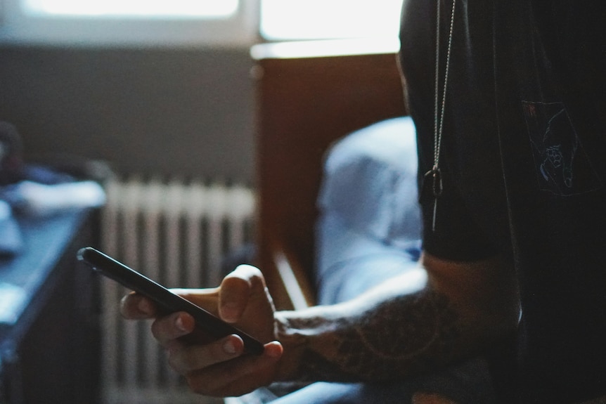 Tattooed arm of man holding phone, sitting on bed.