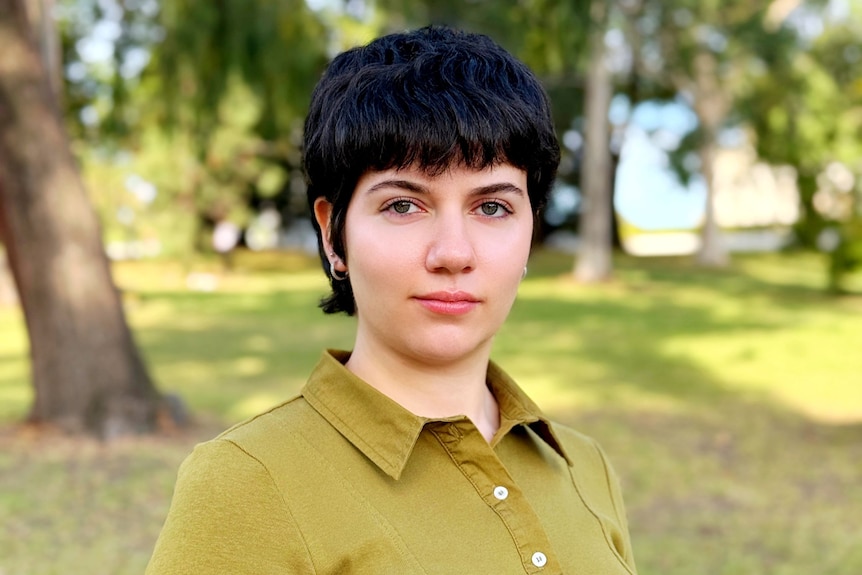 A woman with short black hair, wearing a chartreuse shirt, standing outdoors with gum trees in the background.