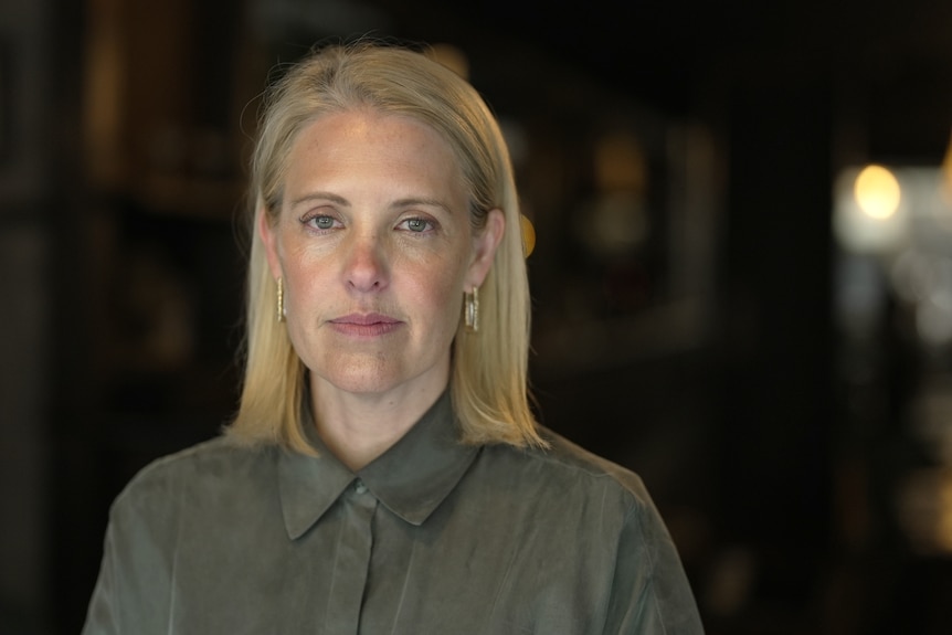 A woman with shoulder-length blonde hair looks into camera with a serious expression.