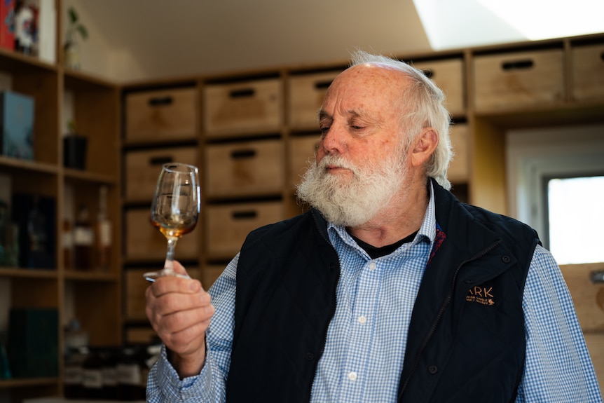 An older man in a blue vest sits in front of whisky barrels in a large shed