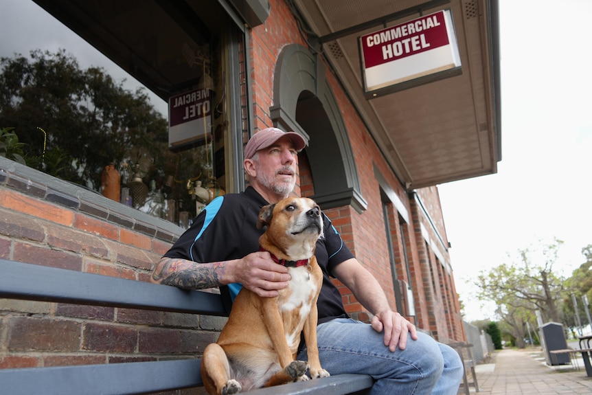 A man and dog sit on a bench outside a building looking towards the town with a sign above them reading: "Commercial Hotel."