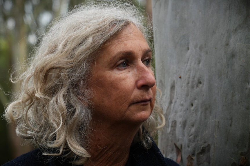 Close up image of Kerry Walker with short hair looks off right of camera in front of tree trunk