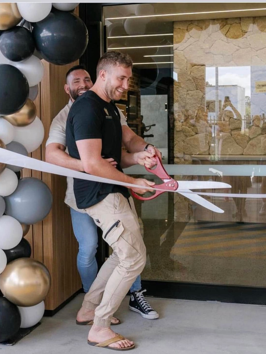 two men embracing each other while cutting ribbon with novelty-sized scissors