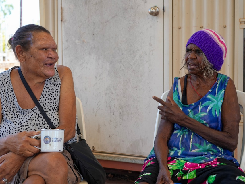 Two Aboriginal female elders sit next to each other gazing at one another warmly.