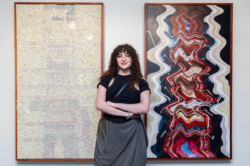 A woman stands in front of two large abstract artworks, one white with text and one rippled in browns and reds