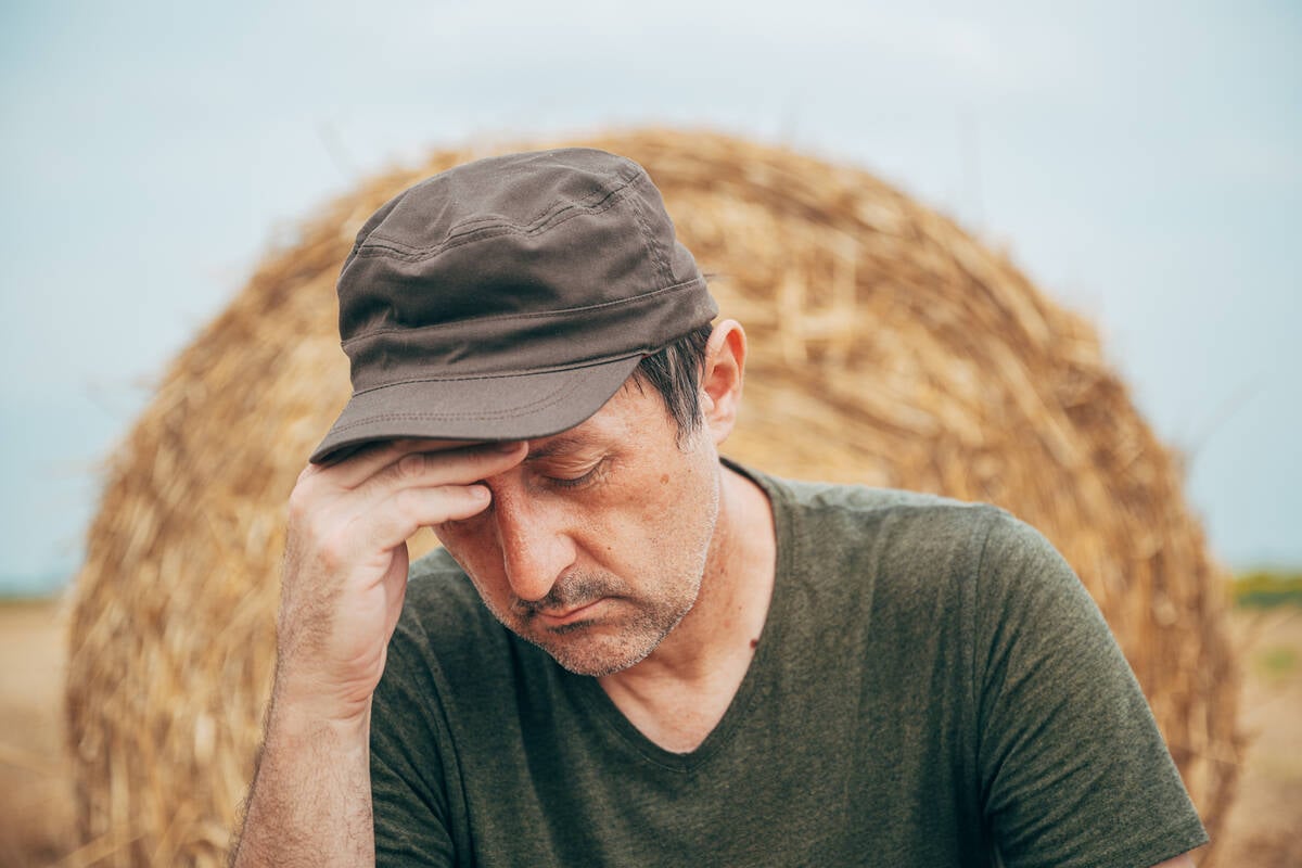 Worried agricultural worker in a cap against a straw bale on a field, reflecting on crop failure or financial loss. Selective focus.