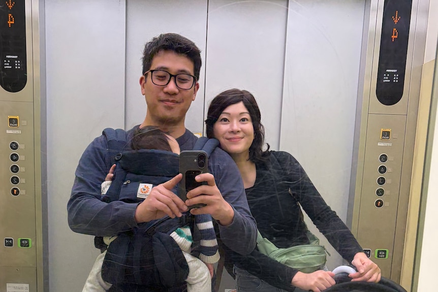 David, carrying his sleeping son in a baby harness, strapped to his chest, while standing next to a woman, his partner.