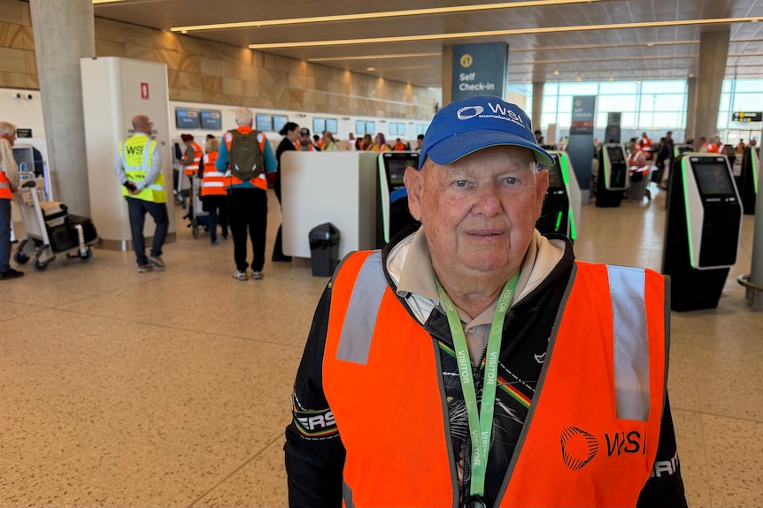 A man stood inside an airport wearing a hi-vis vest and cap.