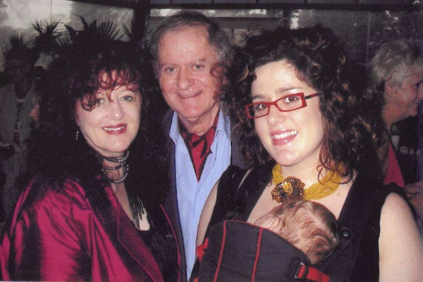 Mother and father with their adult daughter who wears red thick-rimmed glasses and holds a baby in a carrier.