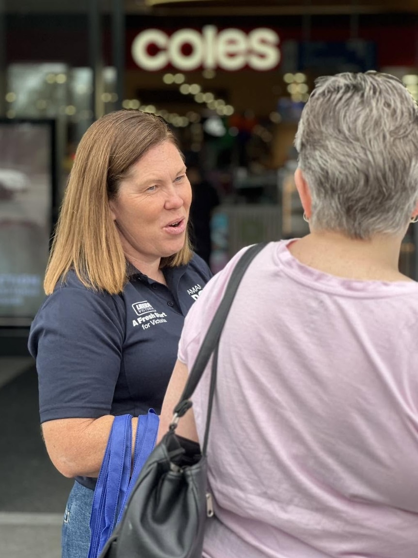 A brown haired woman in a blue collared shirt talks to a grey haired woman in a pink top whose back is to the camera.