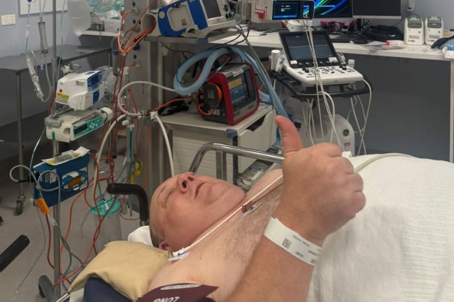 A man lying in a hospital bed gives a thumbs up.