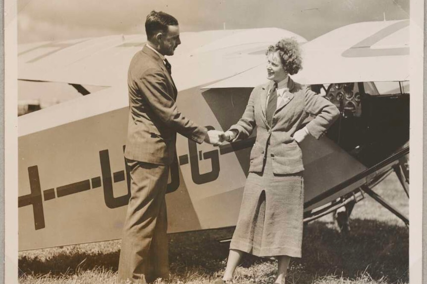 A smiling woman in a skirt suit shakes  hands with a man in a suit as they stand next to aeroplane in a field.