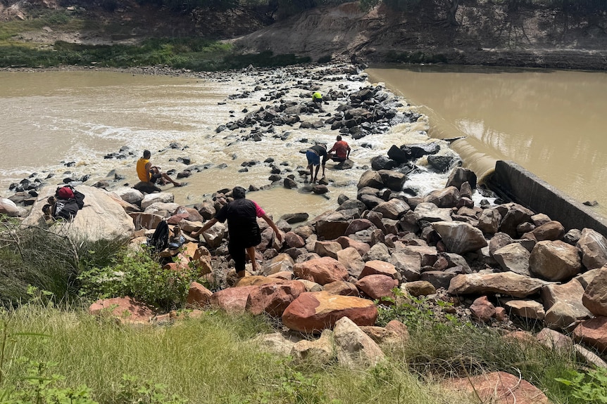 A landscape of a river with a rock weir connecting one bank to the other with five people climbing on the rocks