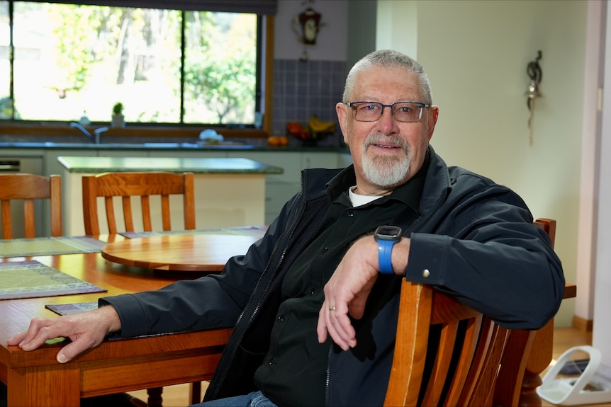 A man with short grey hair and glasses sits at a dining table with one arm slung over the back.