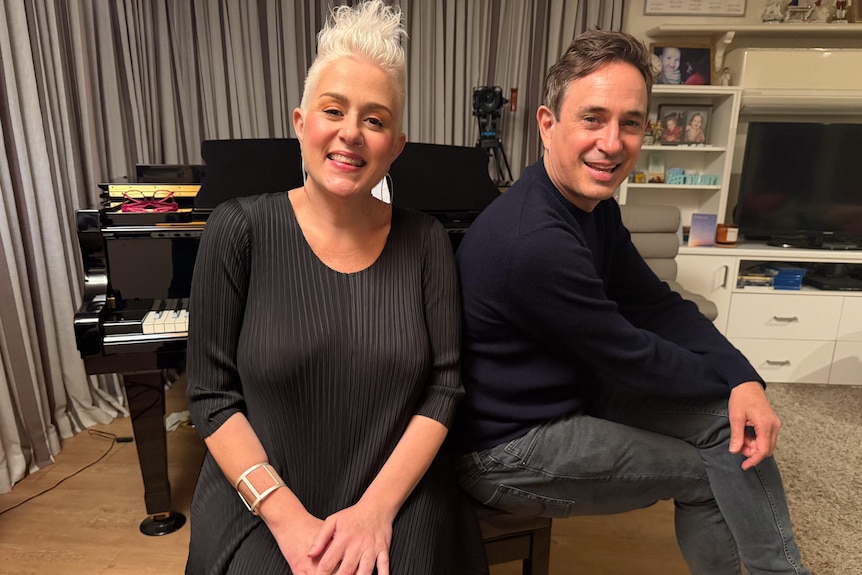 Katie Noonan with short blonde hair wears black, next to Trent Dalton in sweater and long pants smile seated at a piano.