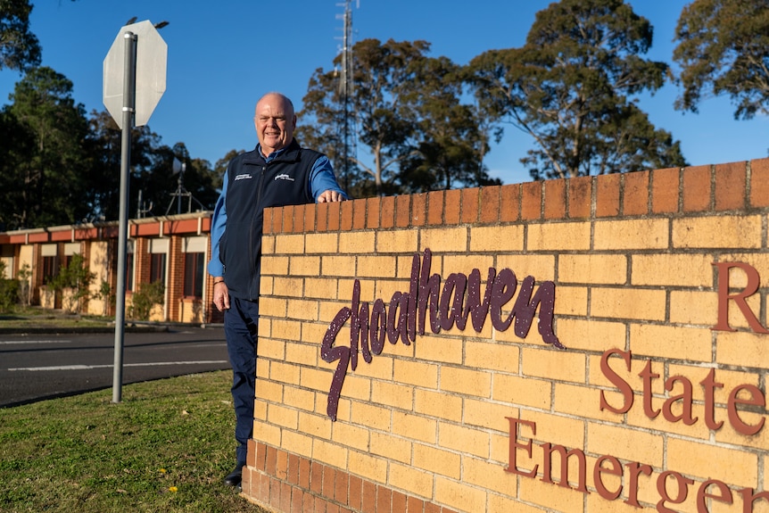 A man in a blue shirt and fleece vest stands next to a brick wall with the sign Shoalhaven State Emergency