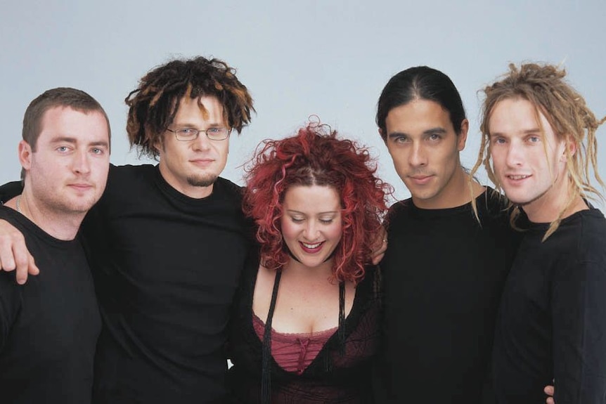 Five people stand close together for a band photo shoot against a plain backdrop.