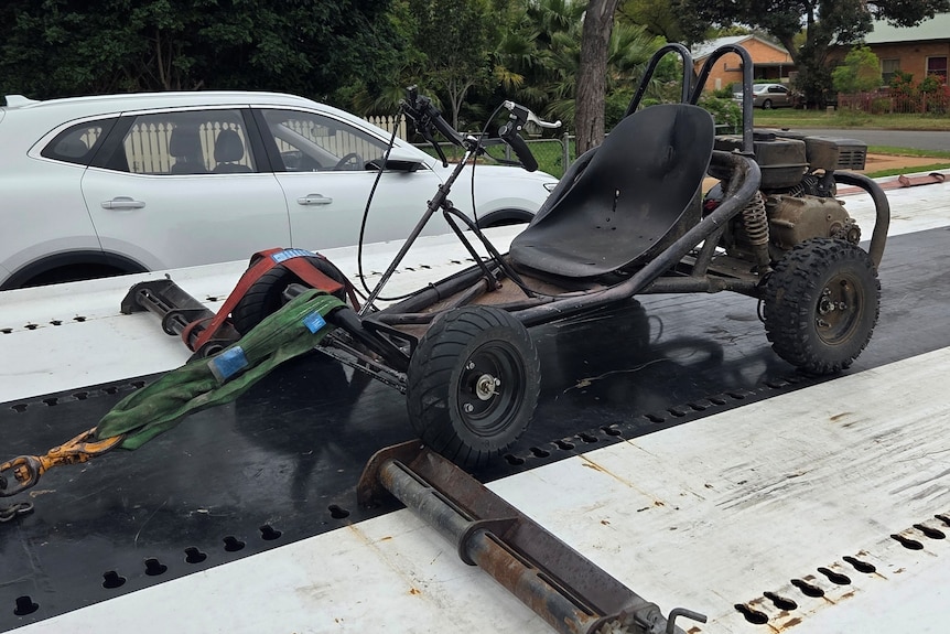 A go-kart is strapped onto the back of a two truck, with a white car in the background.