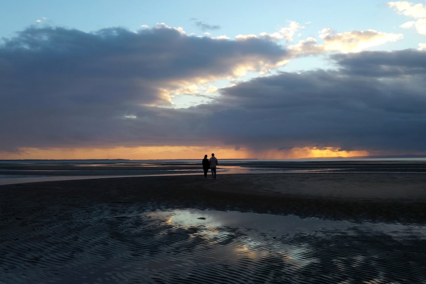 A man and a woman stand in the distance on a beach at sunset, silhouetted.