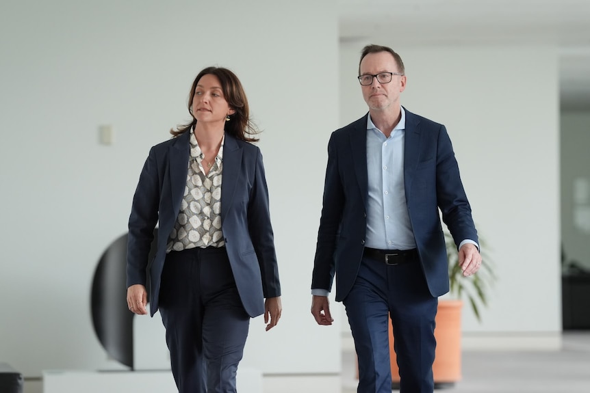A woman and a man, both in suits and walking in a hallway.