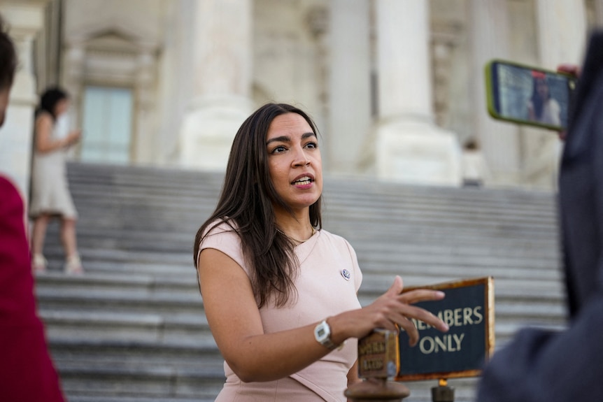 Alexandria Ocasio-Cortez speaks outside the Capitol building. She is in a white dress and has black hair