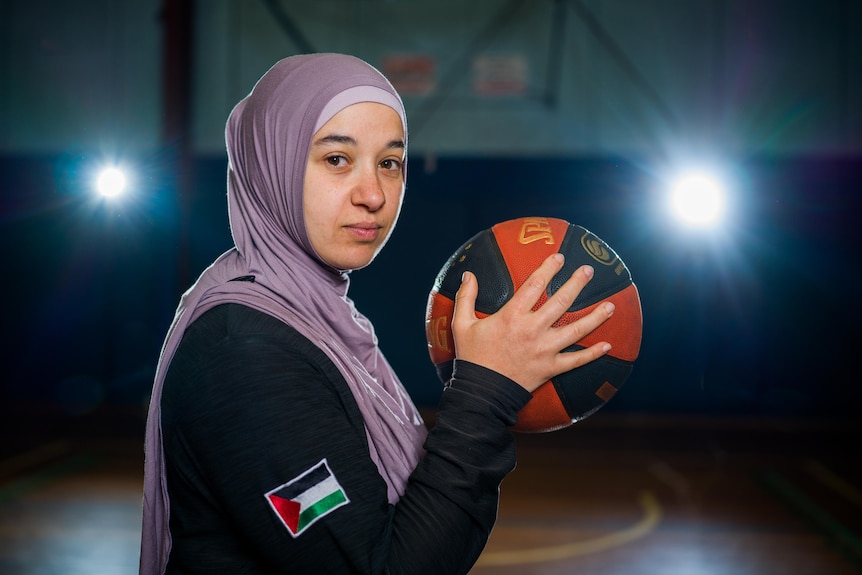 Reema El Qassem, wearing a hijab, looks fiercely at the camera, holding a basketball next to her face.