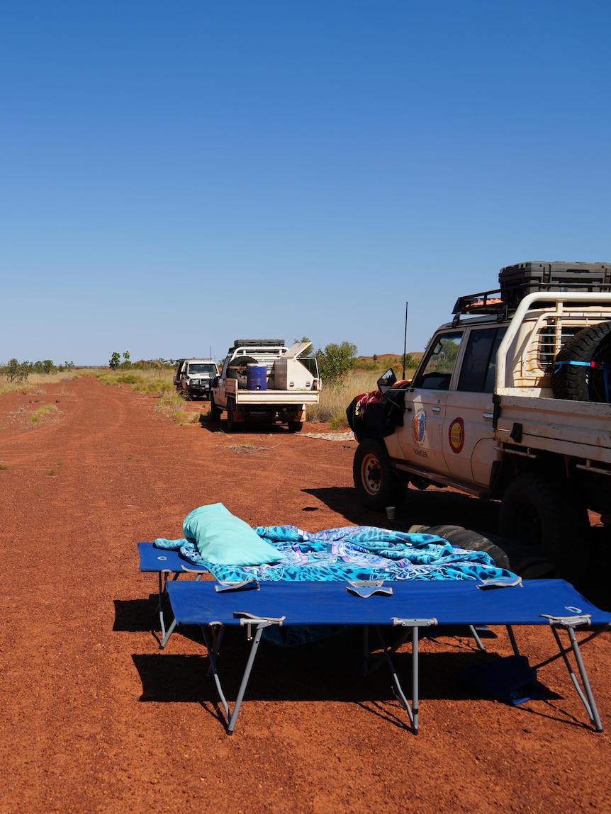 A stretcher camp bed and three four wheel drives line an orange road.