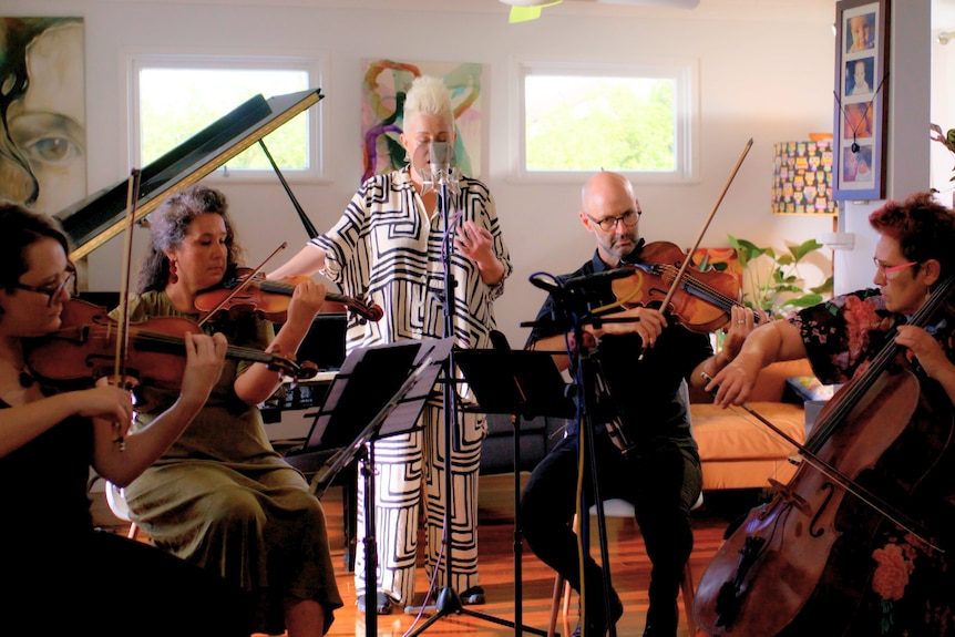 Four-piece string players seated playing in a loungeroom. Katie Noonan stands behind singing into mic.