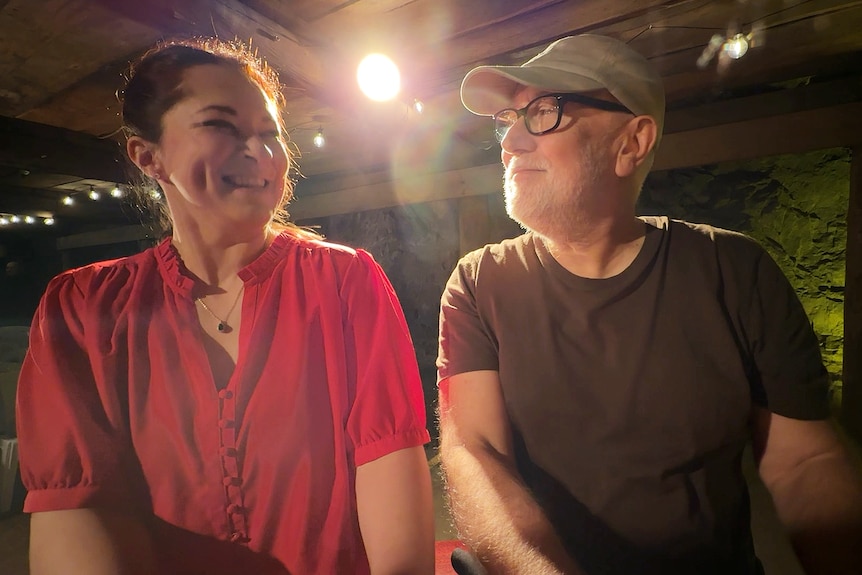 A lady in a red shirt and brown hair and a man in a green t-shirt and baseball cap sit side bu side at a piano