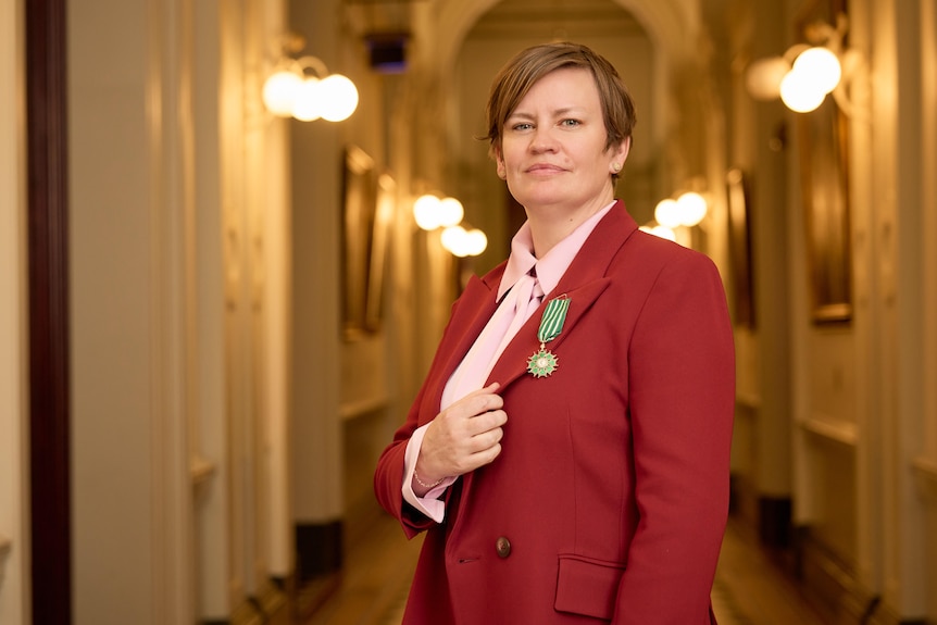 Barbara Moore, a middle-aged woman with short hair and a side fringe, in a red blazer, holds her lapel to highlight a badge.