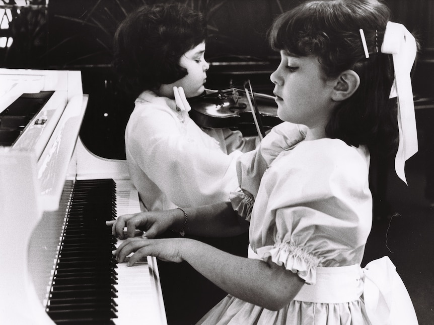 A young girl wearing a dress and ribbon in her hair seated at a piano. Imagery is black and white.
