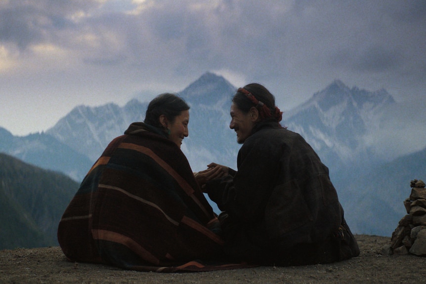 Two people sit under blankets holding hands and looking at each others, with mountains in the background.