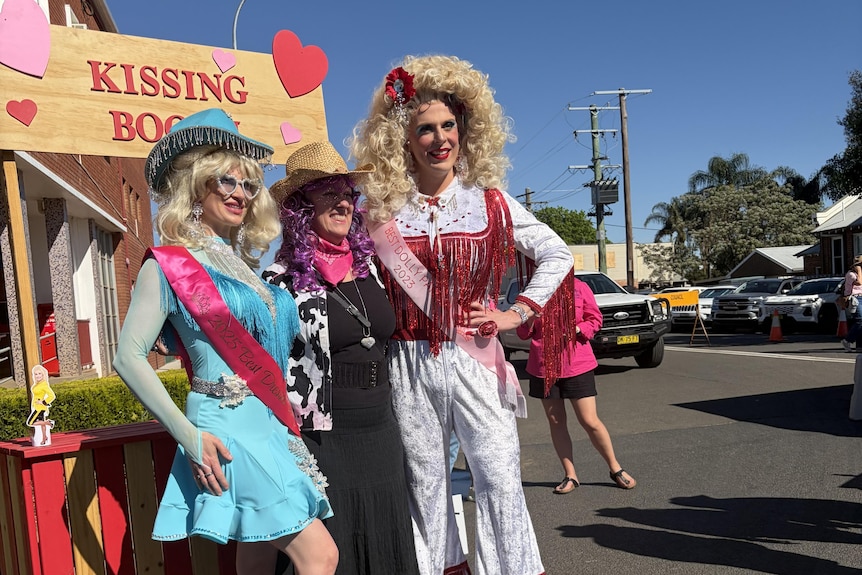 Two people dressed as Dolly Parton with white-haired wigs and colourful costumes pose for a photo with a purple-haired woman.