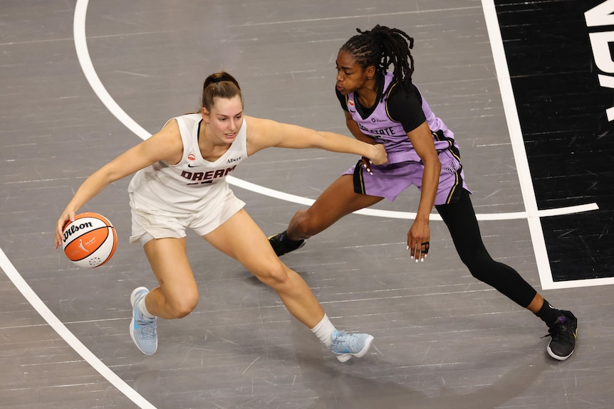 An Australian WNBA player fends off an opposition defender with one hand, holding the ball with the other.