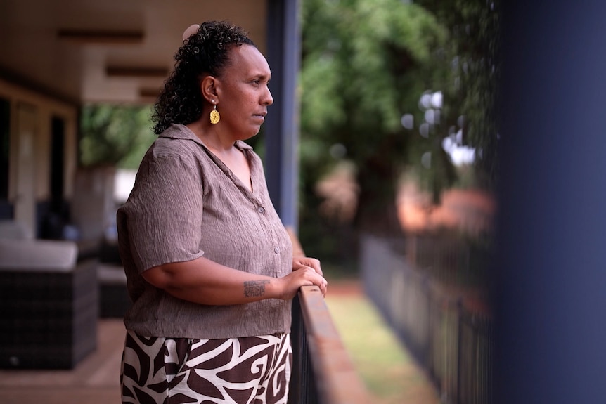 A woman stands on a balcony, holding a railing and looking ahead with a serious expression.