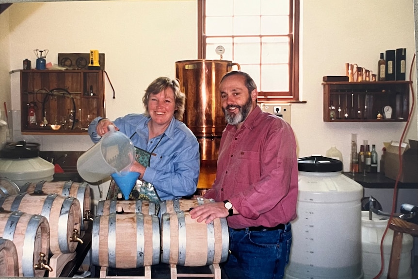 A man and woman standing next to small whisky barrels. The woman is pouring brown liquid through a blue funnel.